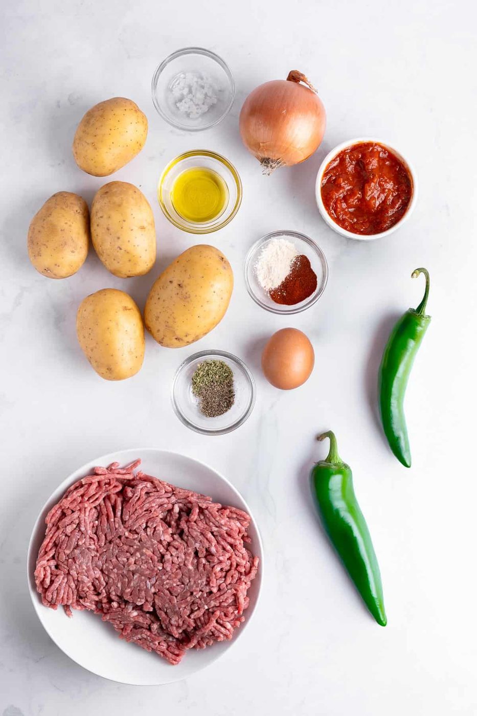 Flat lay of ingredients for a recipe: ground beef in a bowl, several potatoes, two green chili peppers, an onion, small bowls of tomato sauce, flour, spices, olive oil, salt, and a whole egg on a white surface.
