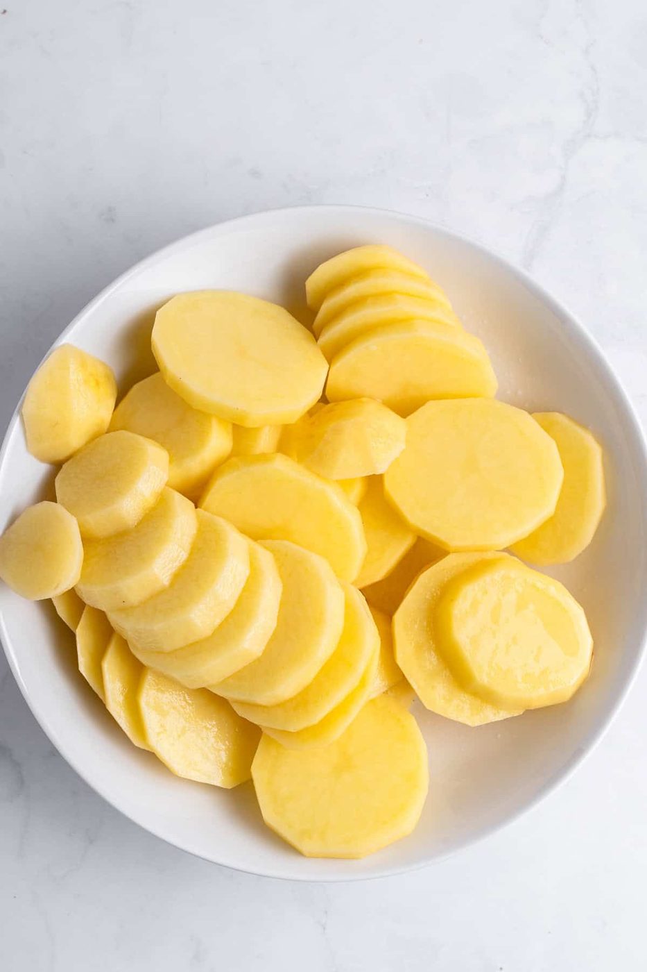 A white bowl filled with thin slices of yellow potatoes on a light marble surface.