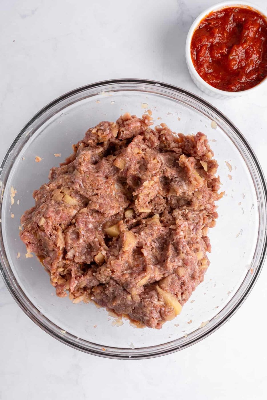 A glass bowl filled with seasoned minced meat on a marble counter. A small white bowl containing red sauce is positioned in the upper right corner.