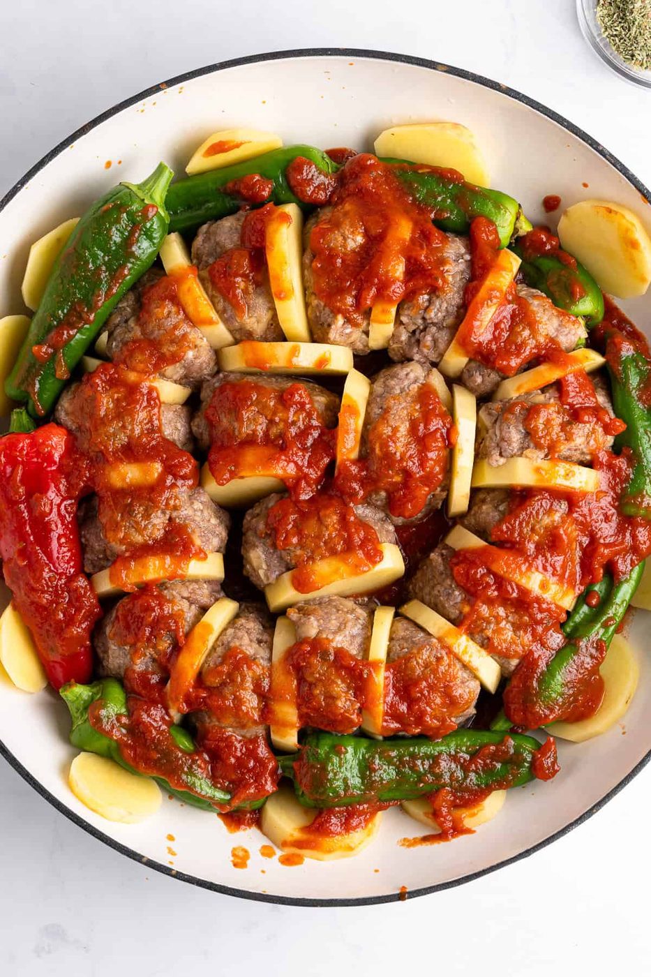 A round dish filled with sliced meat and potato layers, topped with tomato sauce. Surrounded by red and green peppers and additional potato slices. Garnished with herbs.