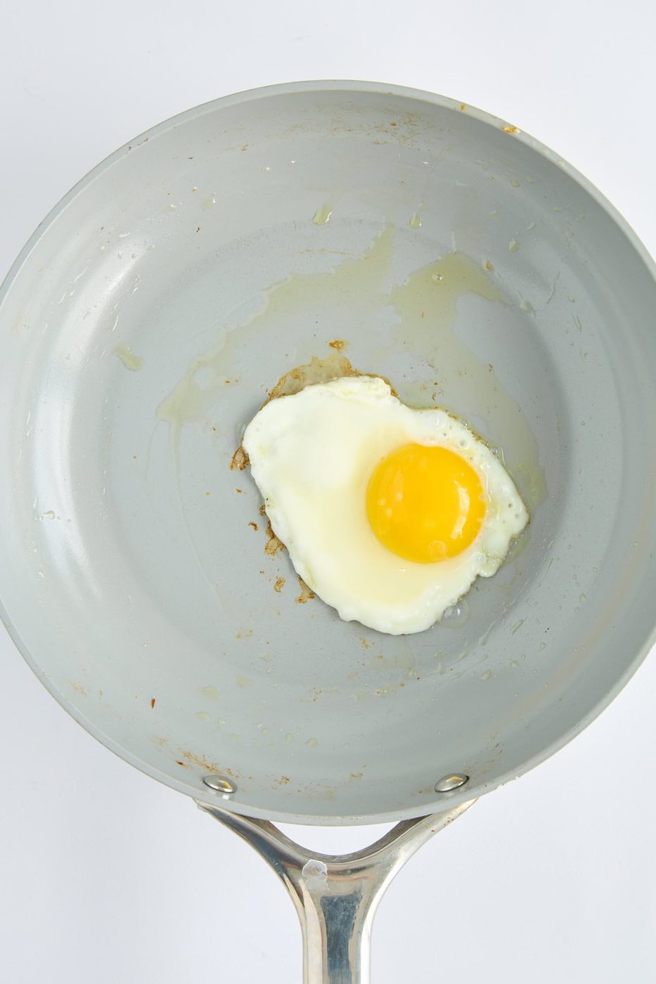 A single sunny-side-up egg frying in a light gray nonstick skillet with a metal handle, seen from above against a white background.