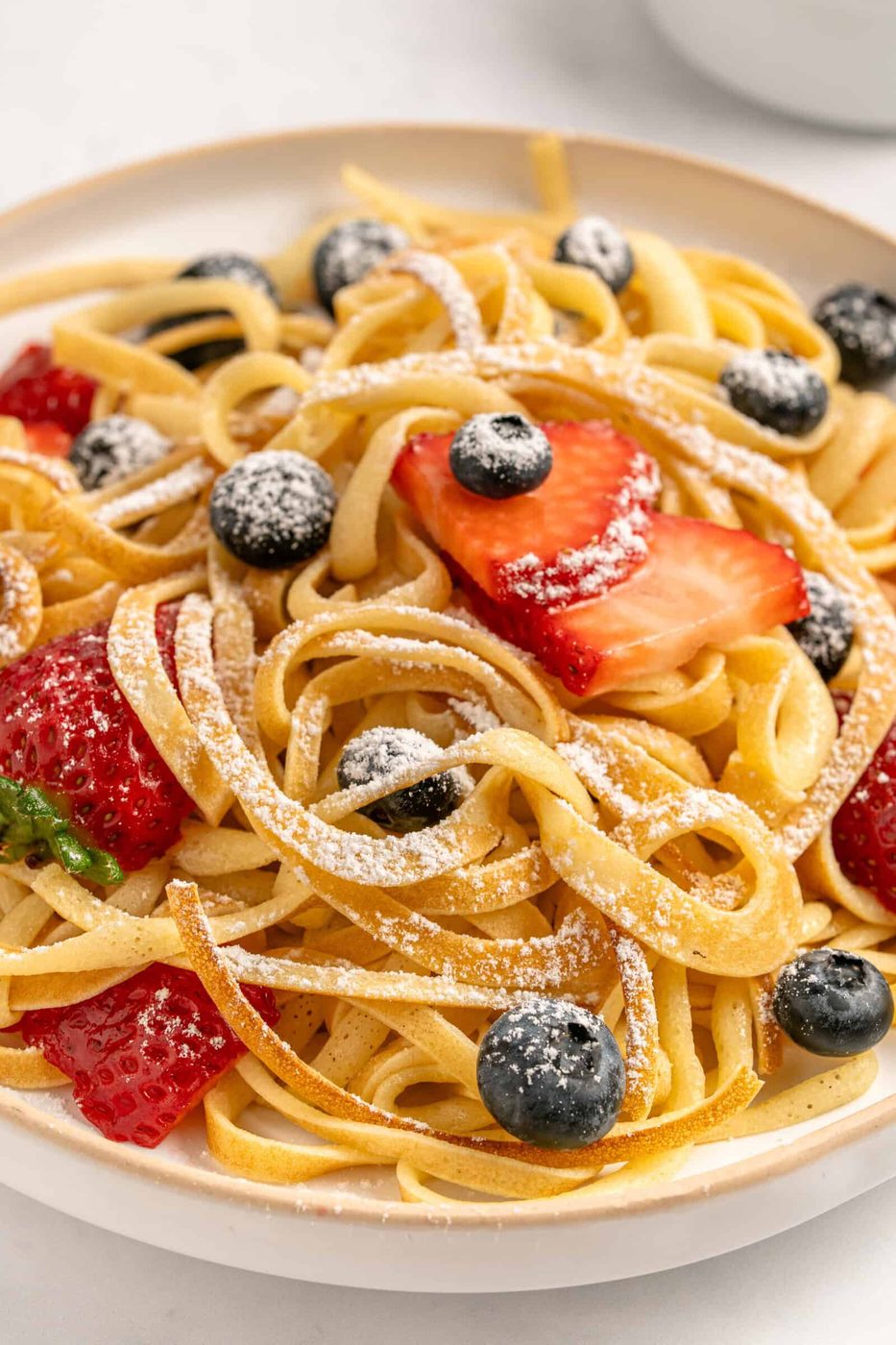 A plate of fettuccine pasta topped with fresh strawberries, blueberries, and a dusting of powdered sugar.