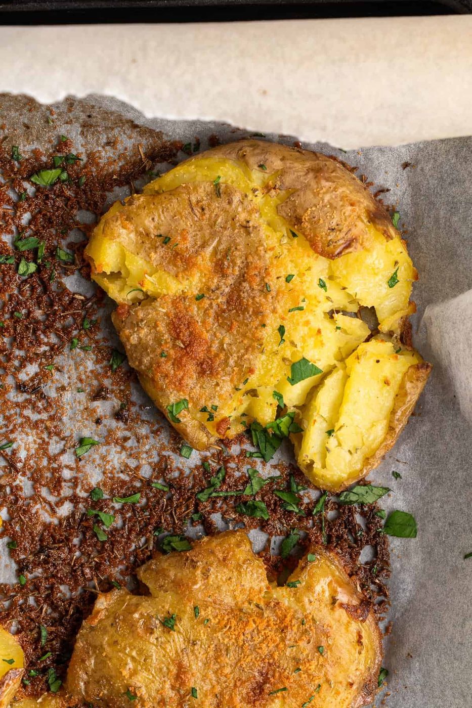 A close-up of crispy potatoes garnished with chopped parsley. The potatoes are golden brown with a textured surface, resting on parchment paper, with bits of seasoning visible around them.