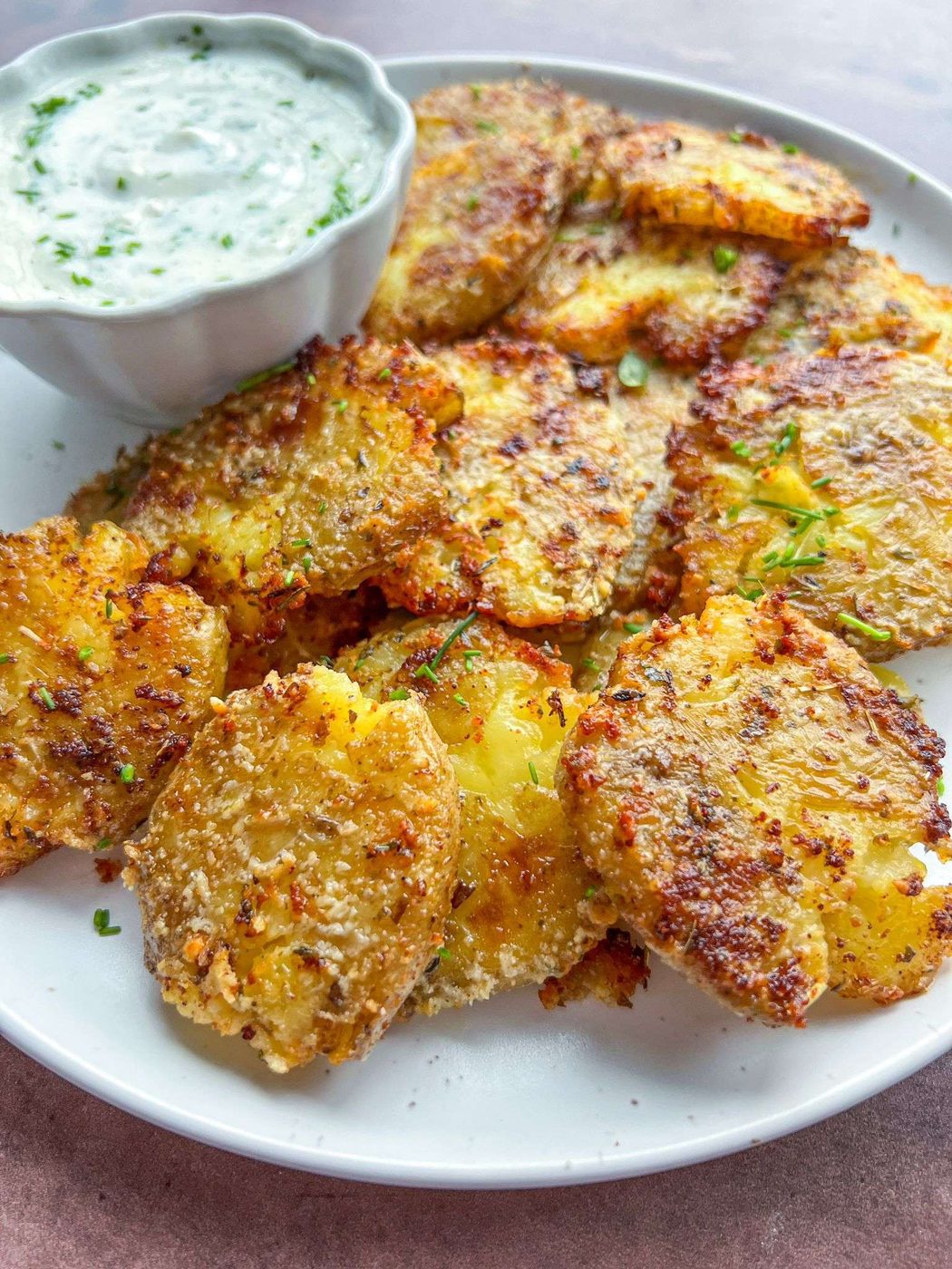 A plate of crispy, golden Parmesan Smashed Potatoes garnished with herbs, served with a small bowl of creamy dipping sauce on the side.