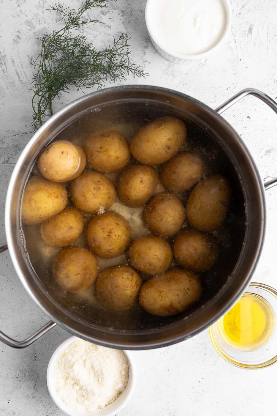 A pot filled with boiling potatoes is centered in the image. Surrounding the pot are small bowls containing butter, flour, and sour cream. Fresh dill is placed nearby on the textured white surface.