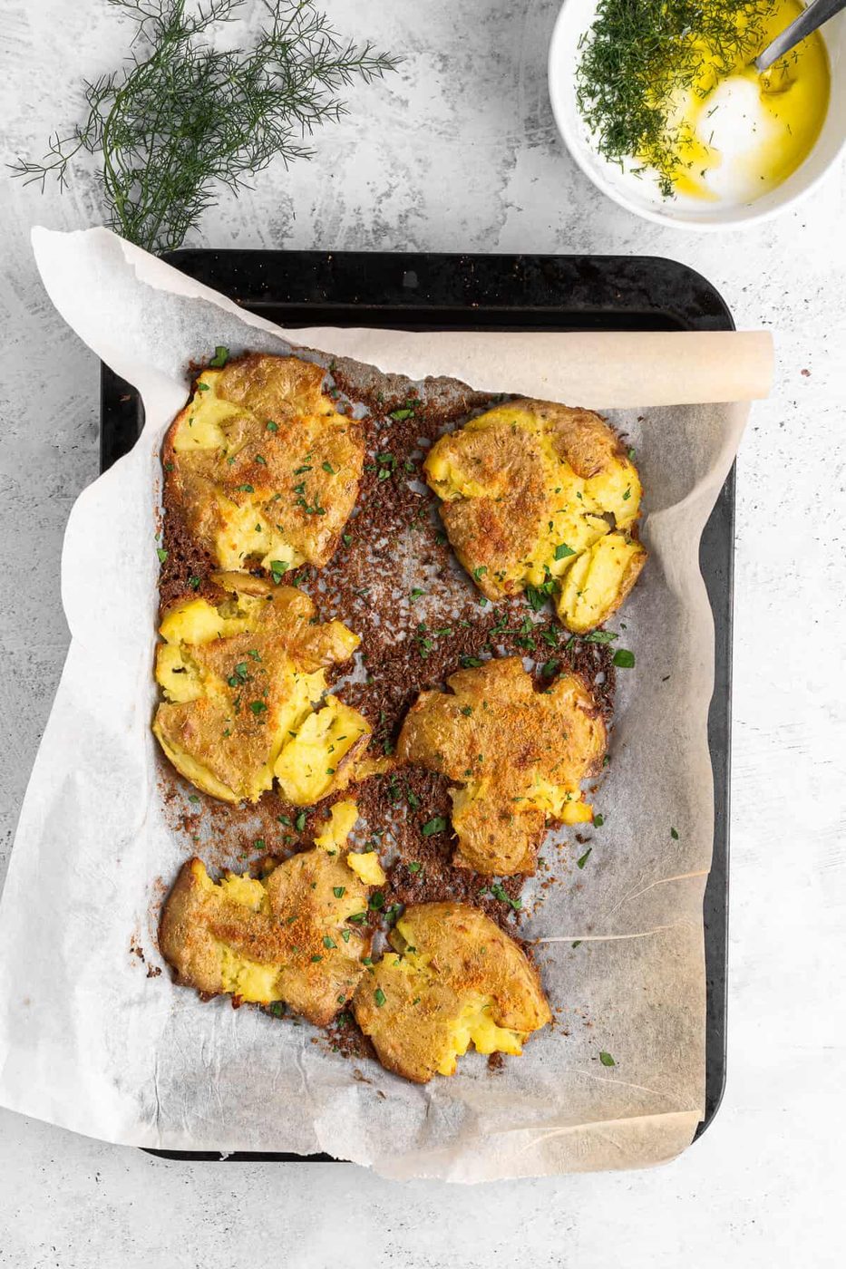 Baked smashed potatoes on a parchment-lined tray, garnished with herbs. A small bowl with olive oil and herbs is in the background. The setting is on a light gray surface with a sprig of fresh herbs nearby.