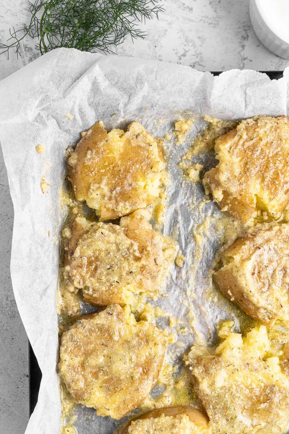 Crushed potatoes on a parchment-lined baking tray, seasoned with herbs and golden-brown baked, with a sprig of fresh dill in the background.