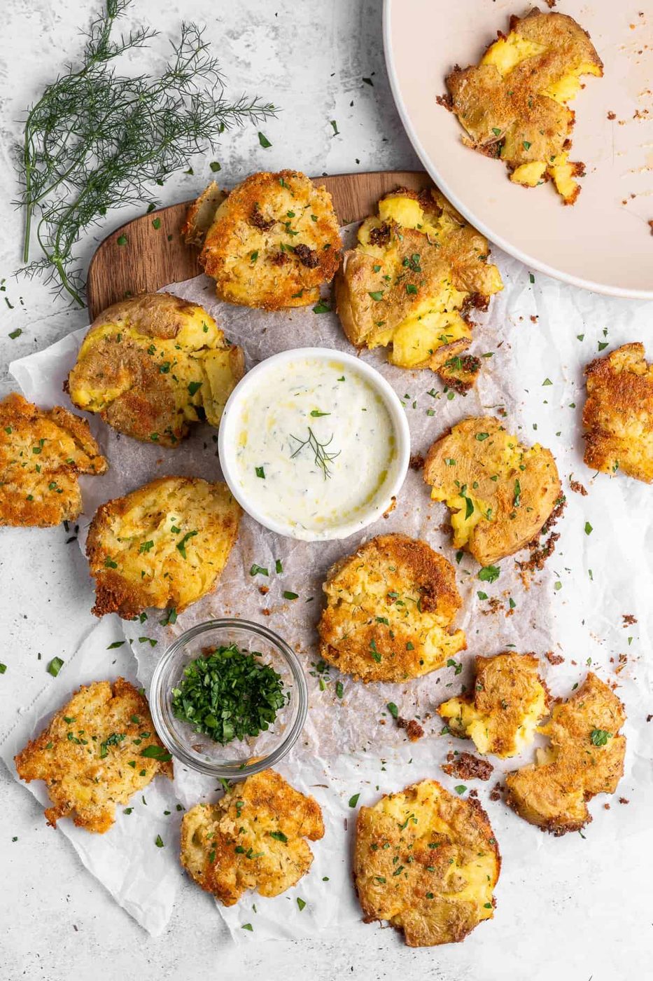 Golden brown smashed potatoes sprinkled with herbs are arranged on parchment paper. A small white bowl of creamy dip garnished with dill is centered on the board. Extra chopped herbs are in a glass jar nearby. A plate with some eaten potatoes is on the side.