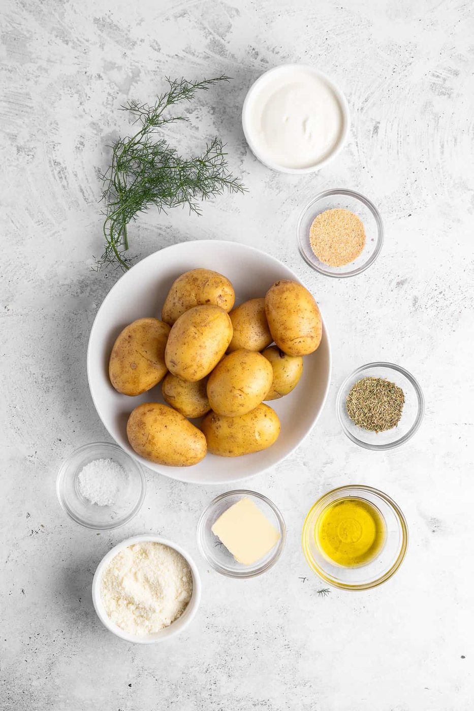 A bowl of potatoes is surrounded by small bowls containing various ingredients: sour cream, dill, garlic powder, dried herbs, olive oil, grated cheese, butter, and salt on a light textured surface.
