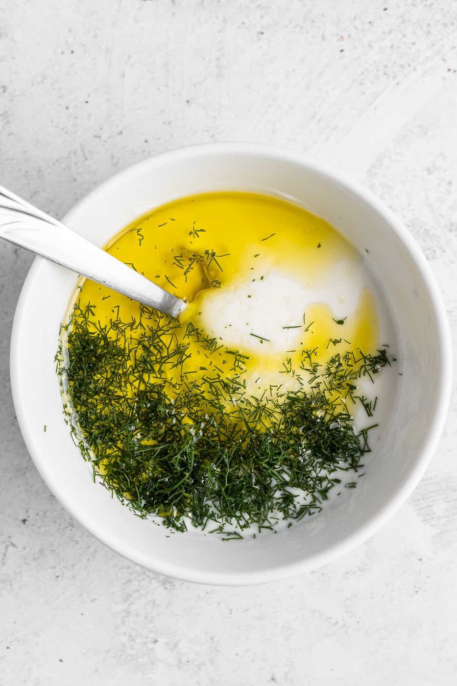 A white bowl containing yogurt, olive oil, and dill, with a spoon resting inside. The ingredients are partially mixed, creating a vibrant contrast of colors and textures. The background is a light, textured surface.