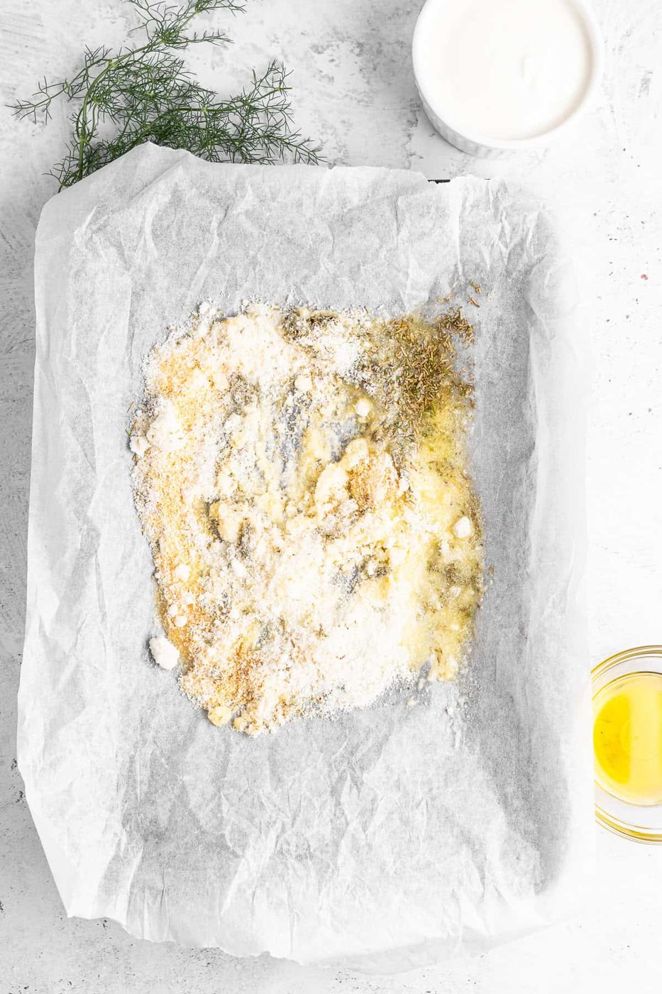 A baking tray lined with parchment paper contains mixed spices, flour, and yogurt, creating a marbled texture. Fresh dill is placed nearby, with a small bowl of yogurt and a glass bowl of olive oil on the side.