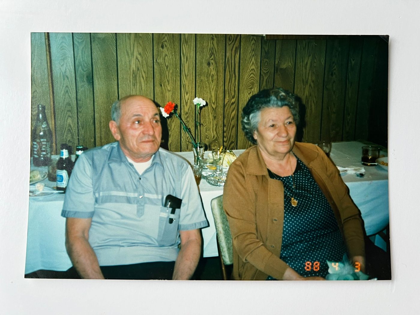 An elderly man and woman sit side by side at a table with empty plates, drinks, and flowers against a wood-paneled wall. Dressed simply and looking relaxed, they might have just enjoyed a comforting bowl of pastina together. The date reads "88 4 3.