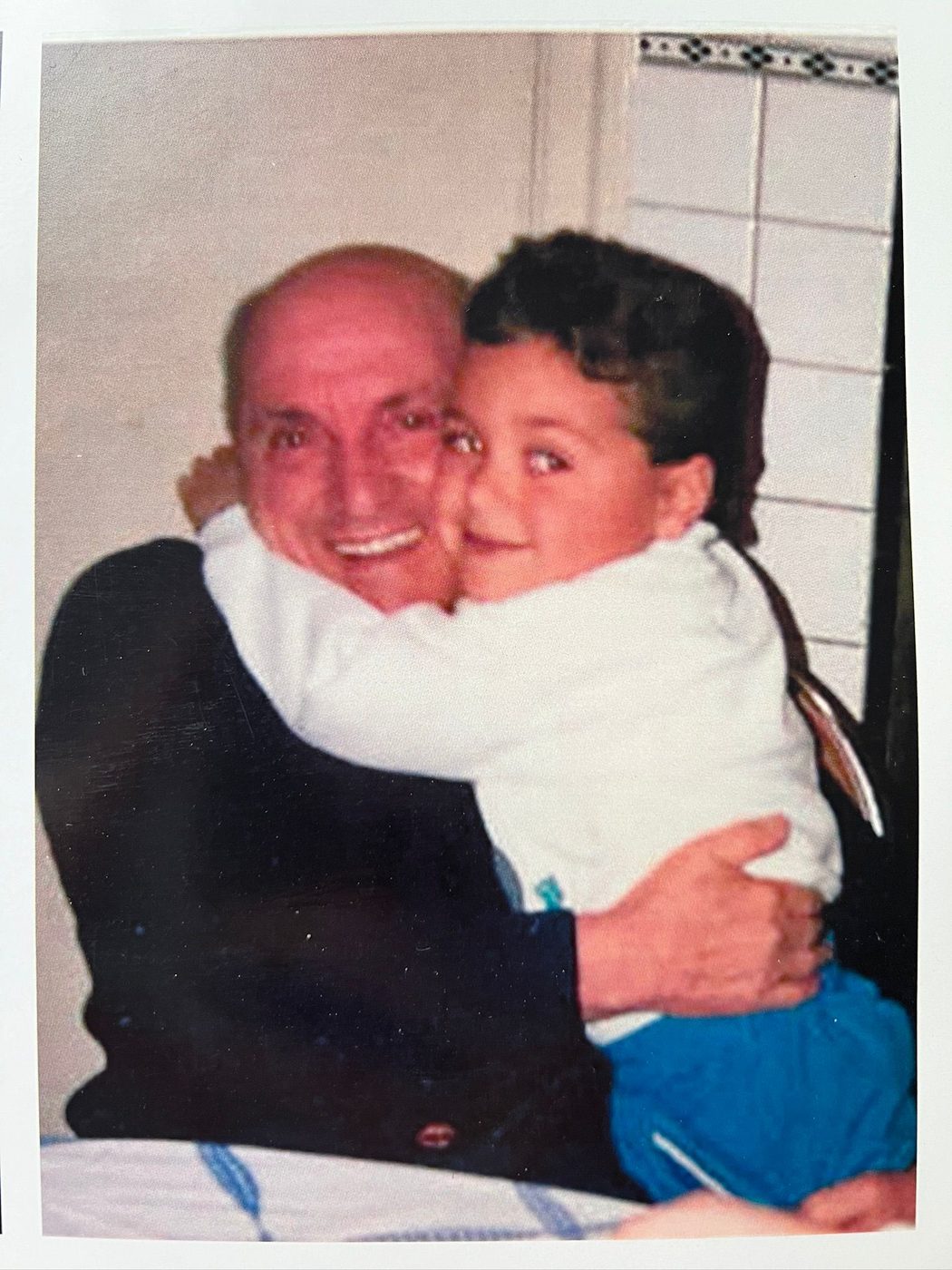A smiling older man and a young boy share a warm hug indoors, radiating happiness and contentment—much like sharing a bowl of comforting pastina together. The man is bald with a dark shirt; the boy has curly hair, white top, and blue pants.