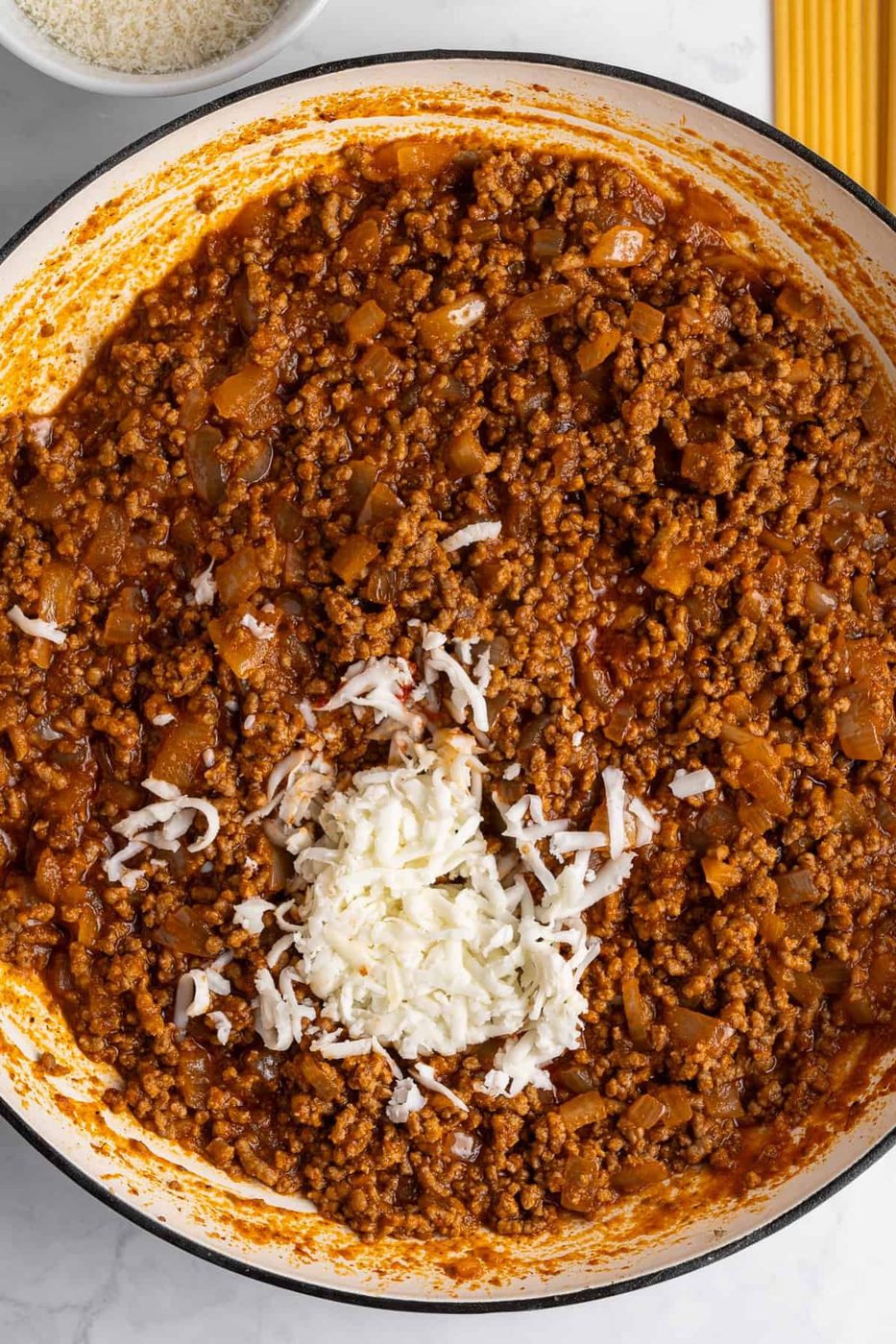 A pot filled with savory bolognese sauce made from ground meat and diced vegetables for the Pastitsio. There's a pile of grated cheese melting in the center. Uncooked spaghetti and a bowl of grated cheese on the side.