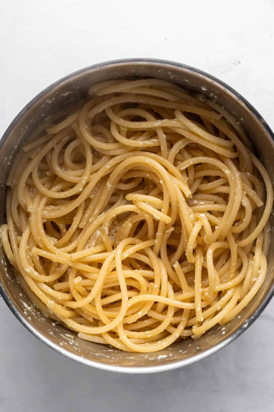 A stainless steel pot filled with spaghetti lightly coated in a creamy parmesan sauce, set on a white marble surface. The pasta is neatly arranged, highlighting the cheese glistening on the strands.