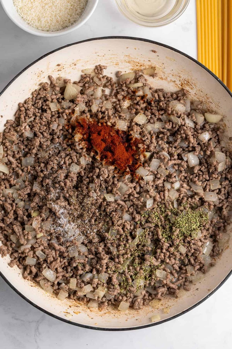 A pan filled with cooked ground beef, chopped onions, and a mix of spices including paprika, black pepper, salt, and herbs. Beside the pan are a bowl of grated cheese, a clear liquid, and uncooked spaghetti on a marble countertop.