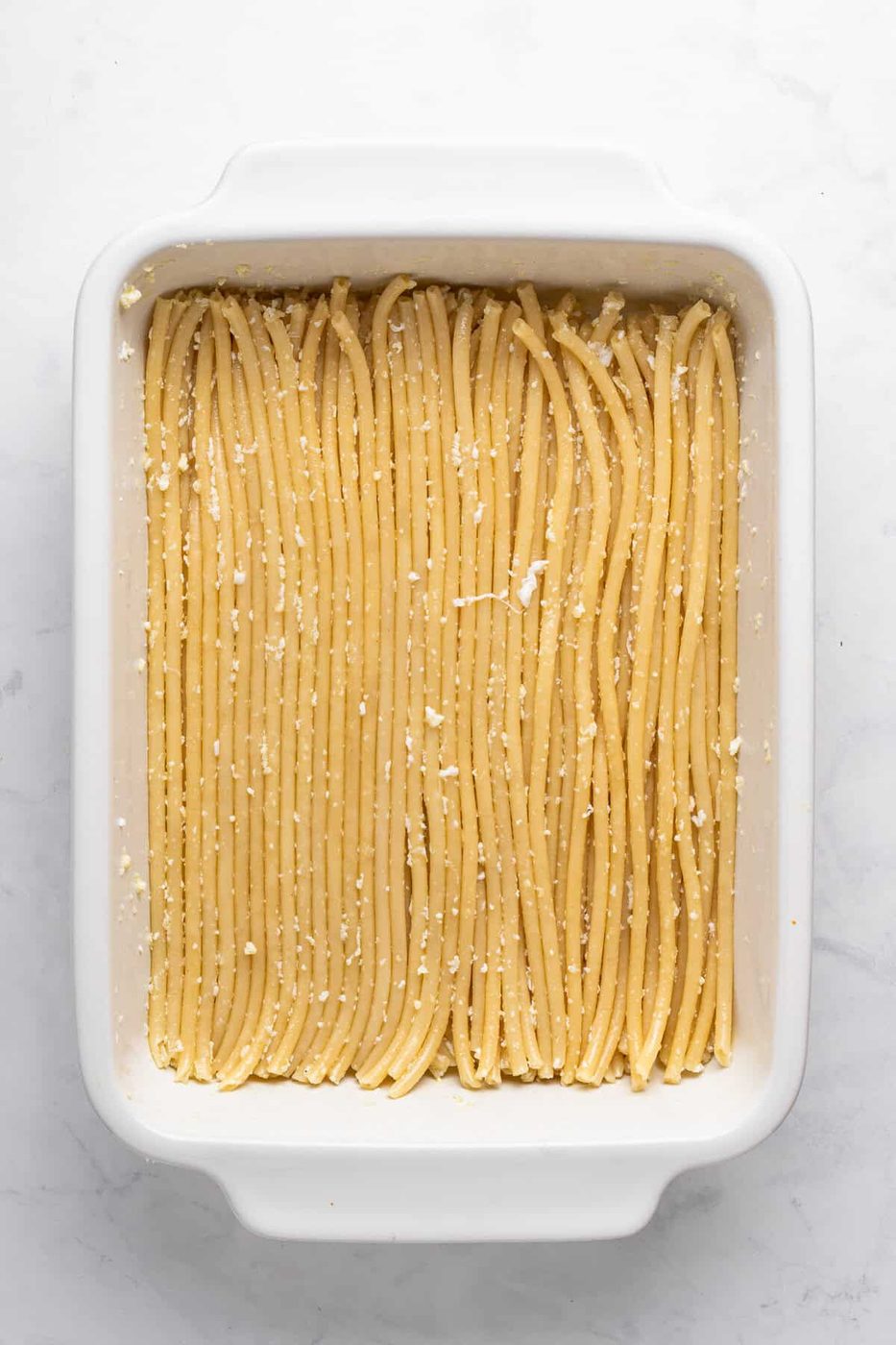 A baking dish filled with neatly arranged, uncooked pasta noodles topped with grated cheese on a white marble countertop.