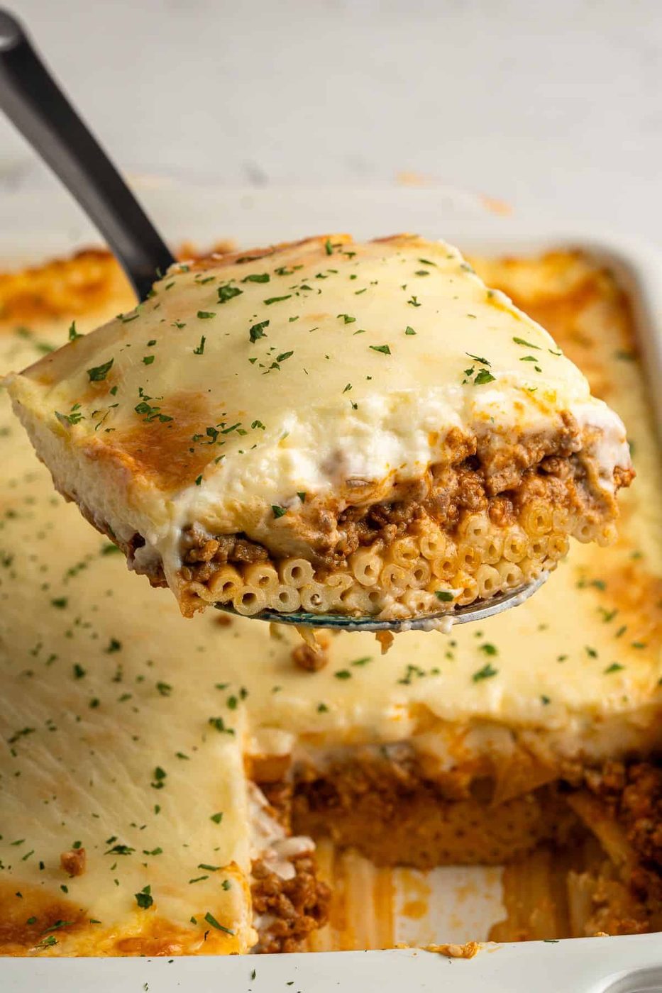 A close-up of a slice of pasta topped with melted cheese and garnish, being lifted from a baking dish. The pasta is layered with a meat sauce and creamy topping, with parsley sprinkled on top. The dish is set against a neutral background.