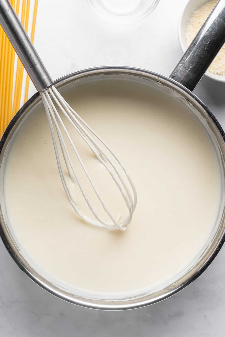 A whisk in a pot of creamy white sauce, likely béchamel, on a light countertop. Uncooked spaghetti and a bowl of breadcrumbs are partially visible nearby.
