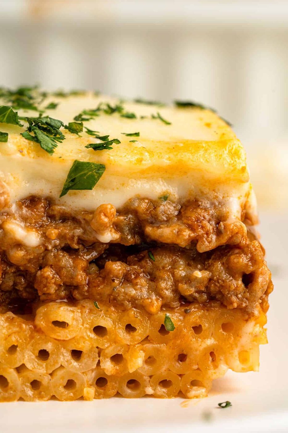 A close-up of a slice of pastitsio, featuring layers of tubular pasta, savory meat sauce, and creamy béchamel on top. Garnished with chopped parsley.