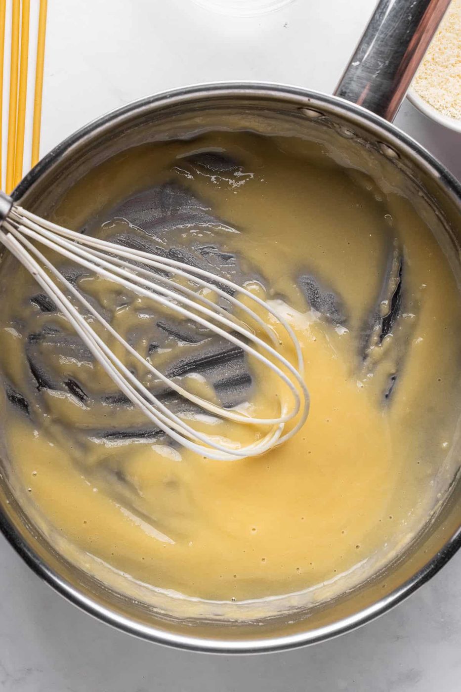 A stainless steel bowl with creamy yellow batter being mixed with a whisk. Spaghetti and a metal pot with breadcrumbs are nearby on a white countertop.