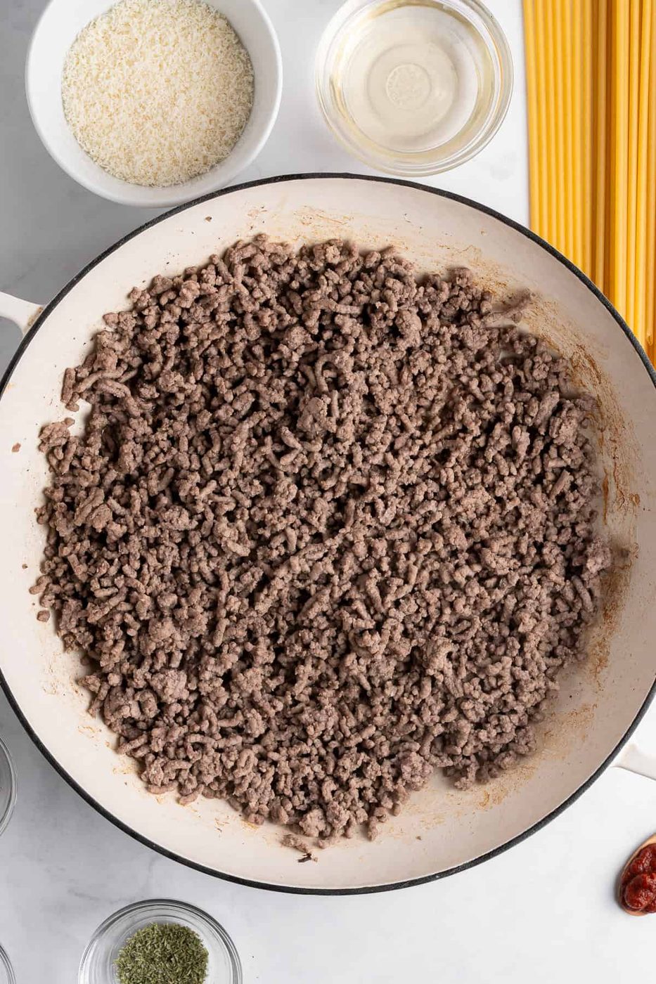 A pan filled with cooked ground meat sits on a countertop. Nearby, uncooked spaghetti, a bowl of breadcrumbs, a small glass bowl of oil, and various spices are positioned, ready for meal preparation.