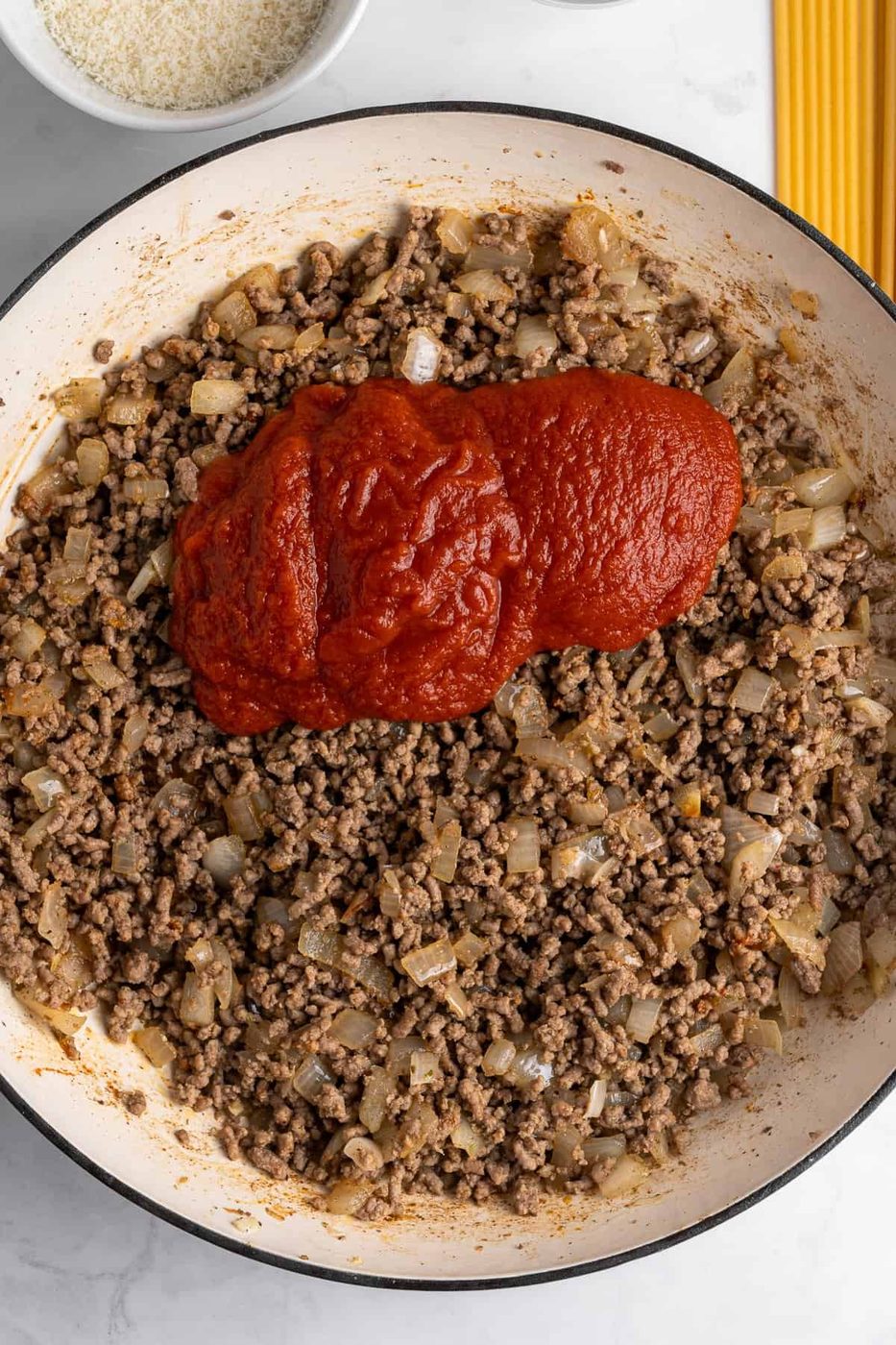 A skillet containing cooked ground beef and diced onions, topped with a generous amount of tomato paste. In the background, there's a bowl of grated cheese and uncooked spaghetti on a marble surface.