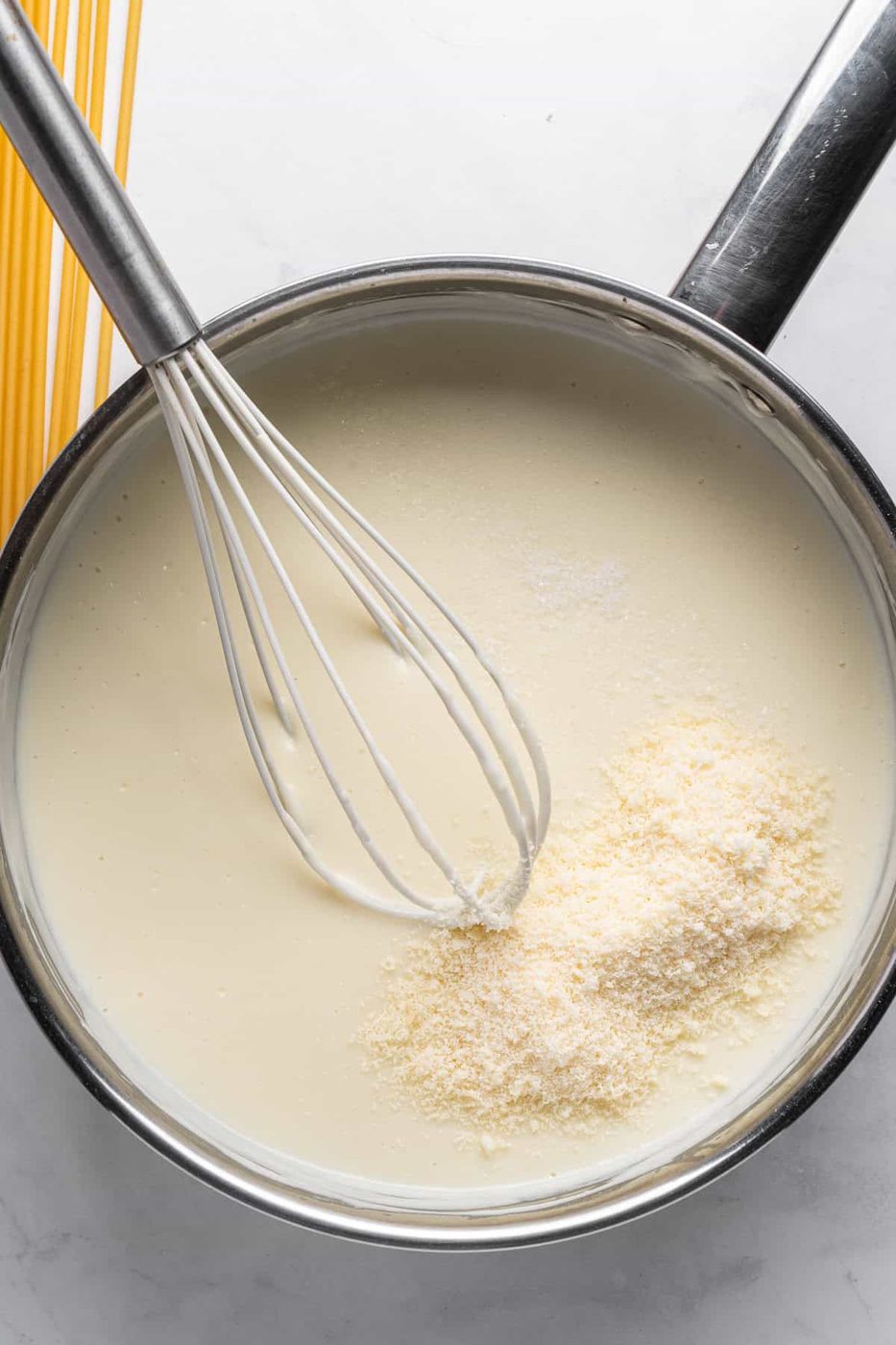 A saucepan filled with white sauce being whisked, with a pile of grated cheese on top for the Pastitsio. Uncooked spaghetti is visible in the background on the countertop.