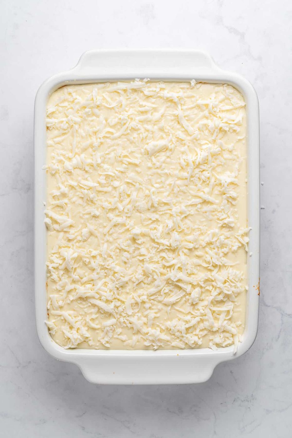 A rectangular white baking dish filled with a Pastitsio topped with a layer of shredded coconut. The dish is set against a light gray marble surface.