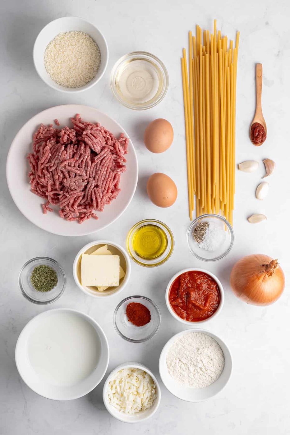 A flat lay of Pastitsio ingredients on a white surface: ground meat, dry spaghetti, eggs, onion, garlic cloves, tomato paste, tomato sauce, olive oil, butter, flour, milk, cheese, spices, and breadcrumbs in various bowls and containers.