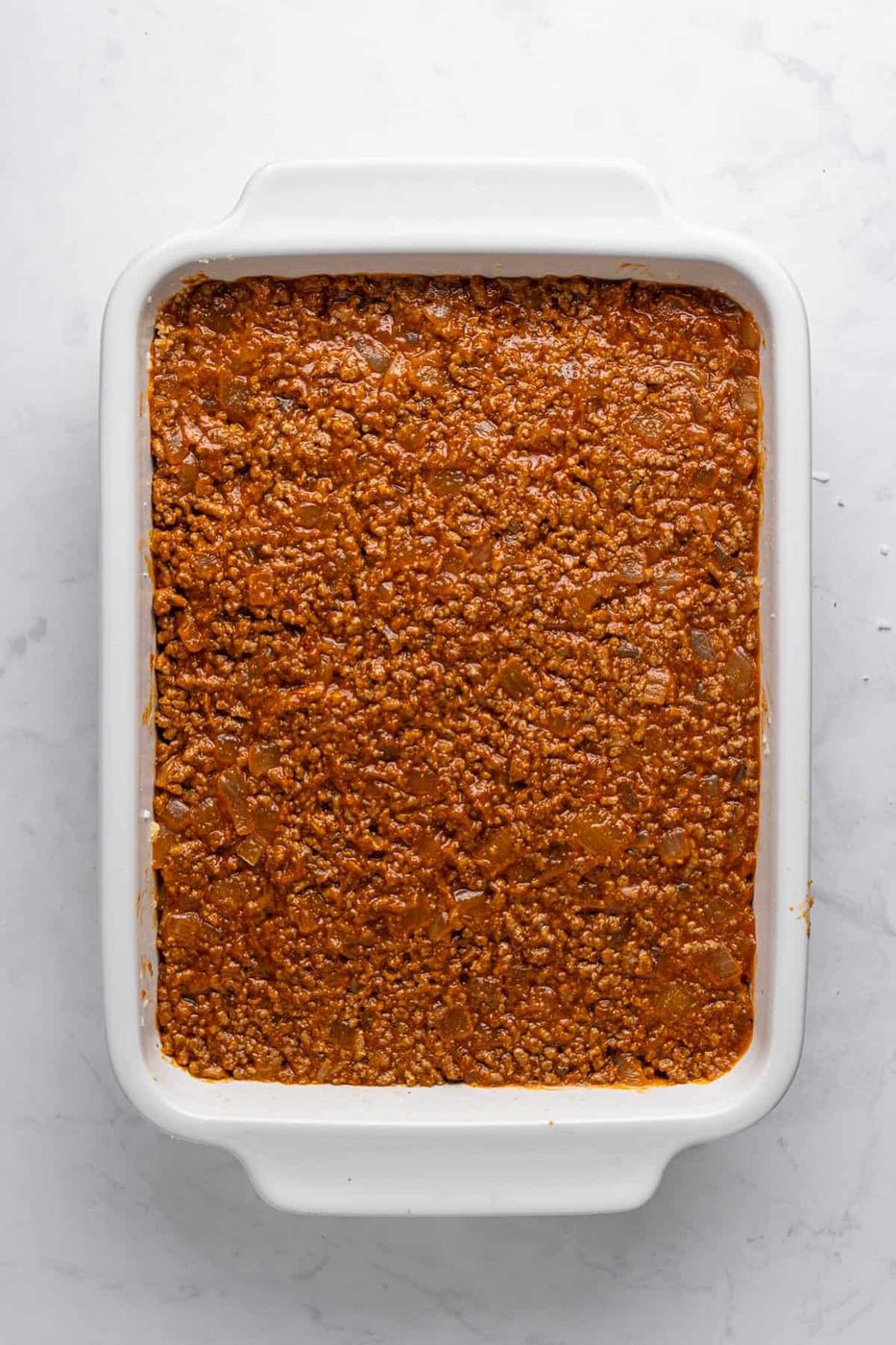 A white rectangular baking dish filled with a layer of finely ground meat sauce, cooked to a rich brown color, is placed on a light gray marble surface.