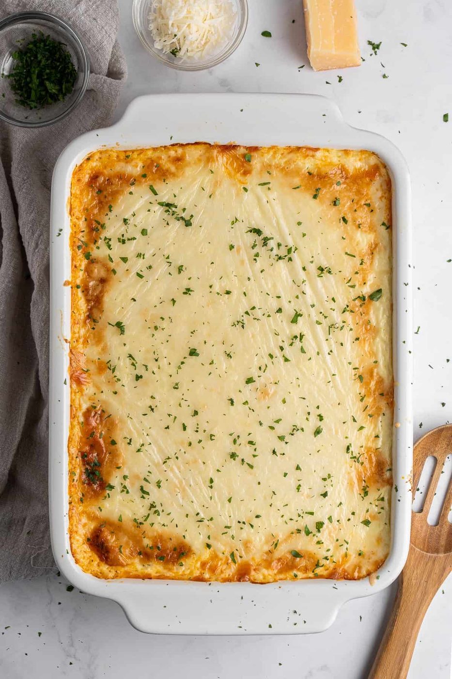 A golden-brown baked Pastitsio in a white rectangular dish, garnished with chopped parsley. Surrounding the dish are bowls of grated cheese and chopped herbs, a block of cheese, and a wooden spatula on a light-colored countertop.