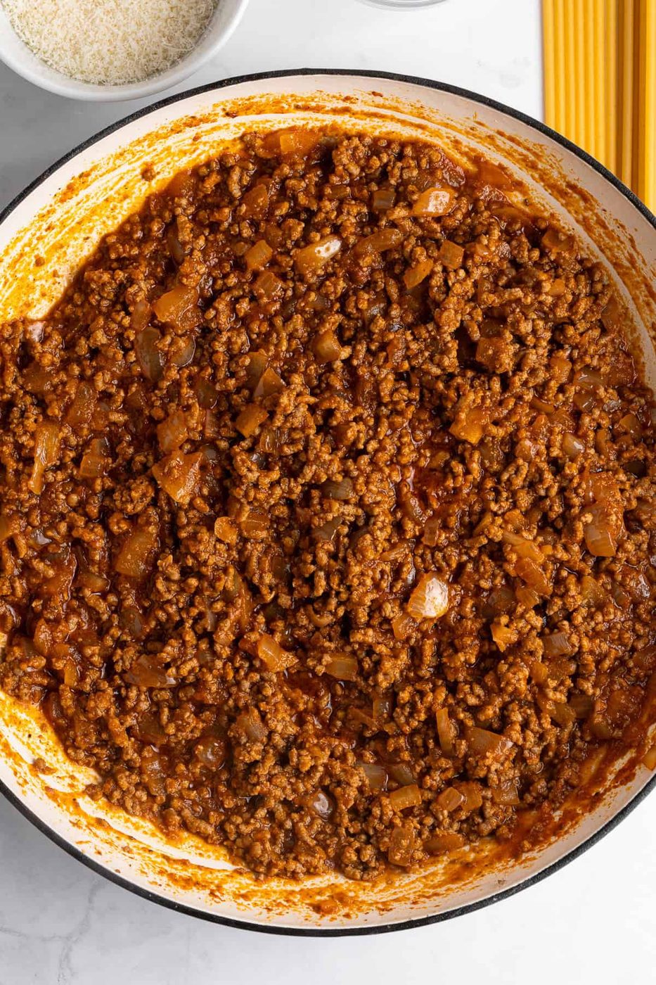A close-up of a pan filled with rich red meat sauce, containing ground meat and visible bits of onion, with a small plate of grated cheese and uncooked spaghetti nearby. The sauce appears thick and well-cooked.