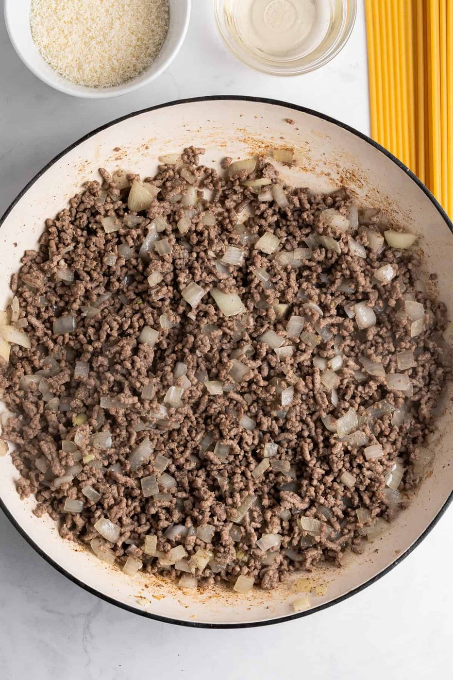 A large pan contains cooked ground beef mixed with diced onions. Nearby are uncooked spaghetti strands, a small bowl of grated cheese, and a glass bowl of clear liquid on a light marble surface.
