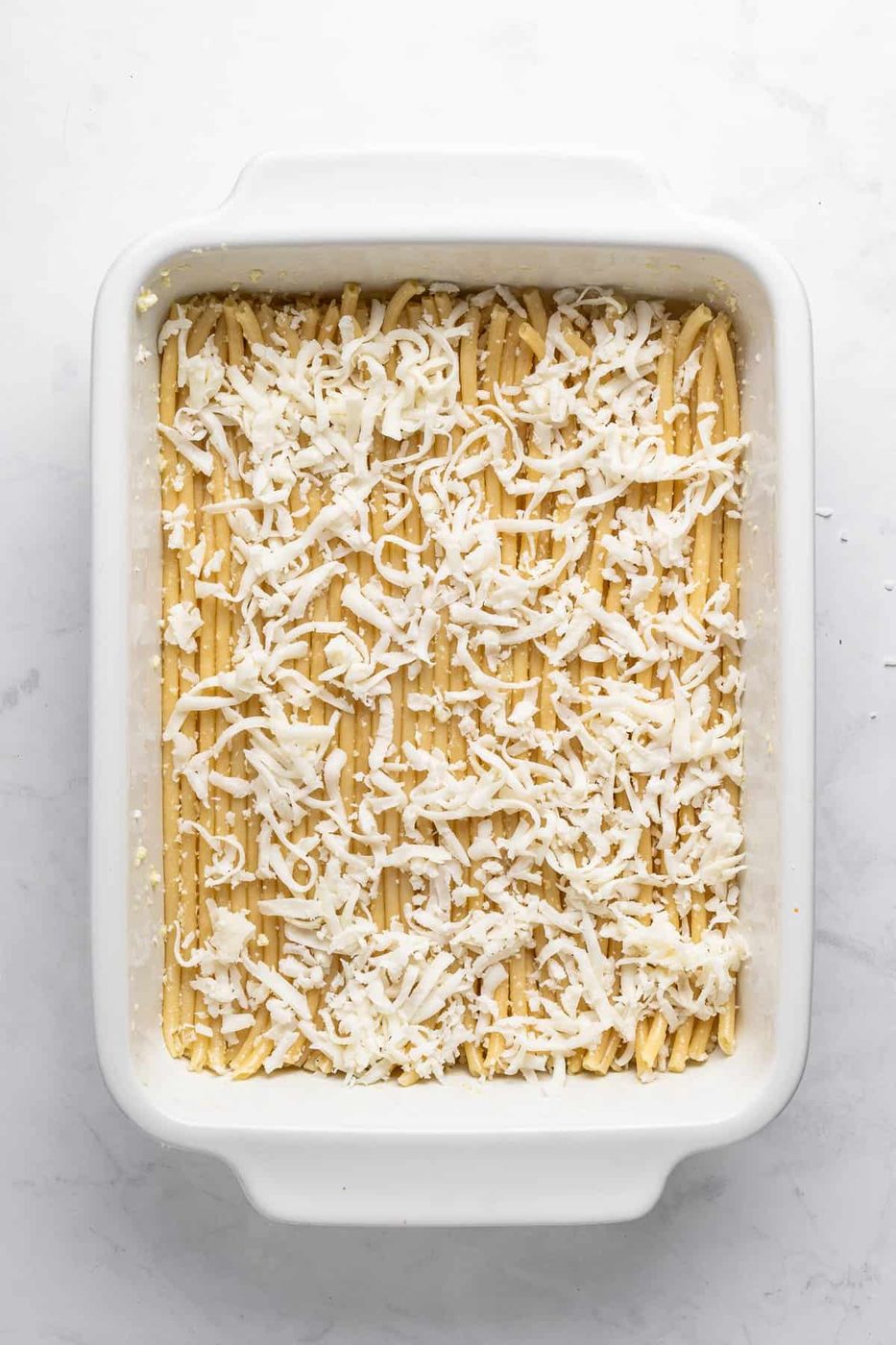 A white rectangular baking dish filled with lasagna noodles covered in shredded cheese, ready for baking. The background is a marble countertop.