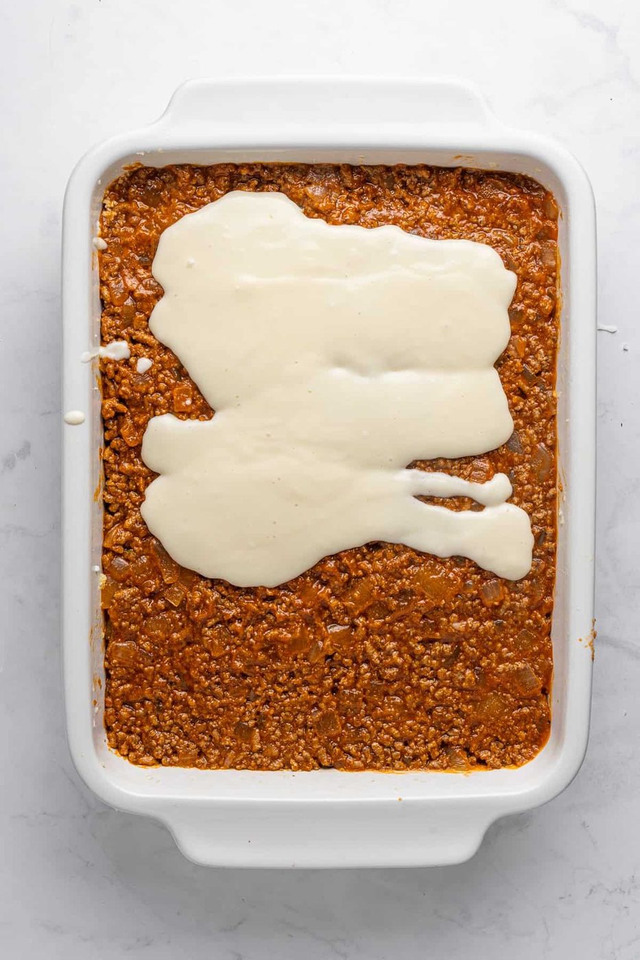 A rectangular white baking dish filled with a layer of cooked minced meat topped with a light tan sauce, spread unevenly. The dish sits on a marbled white countertop.
