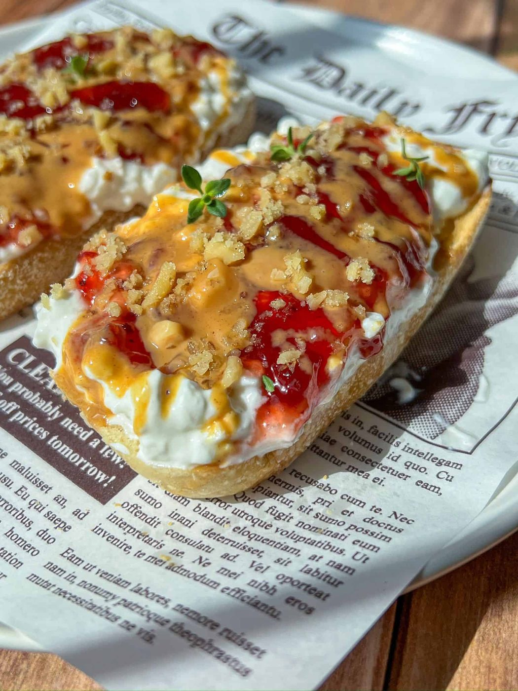 Two slices of bread topped with creamy cheese, red jam, a drizzle of caramel or honey, crunchy crumbs, and fresh herbs, served on a plate lined with newspaper-style paper.