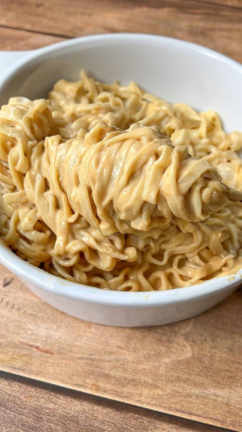 A close-up of creamy, cooked peanut noodles in a white bowl, with a fork lifting a portion. This inviting Peanut Noodles is served on a wooden surface, ready to enjoy.