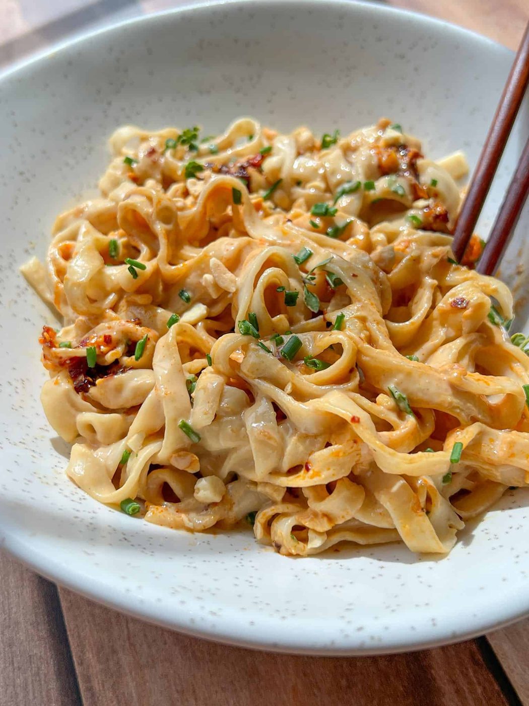 A bowl of wide, flat noodles tossed in a creamy, spicy sauce, garnished with chopped chives. Chopsticks rest on the edge of the bowl. The noodles are served on a light-colored, speckled ceramic dish.