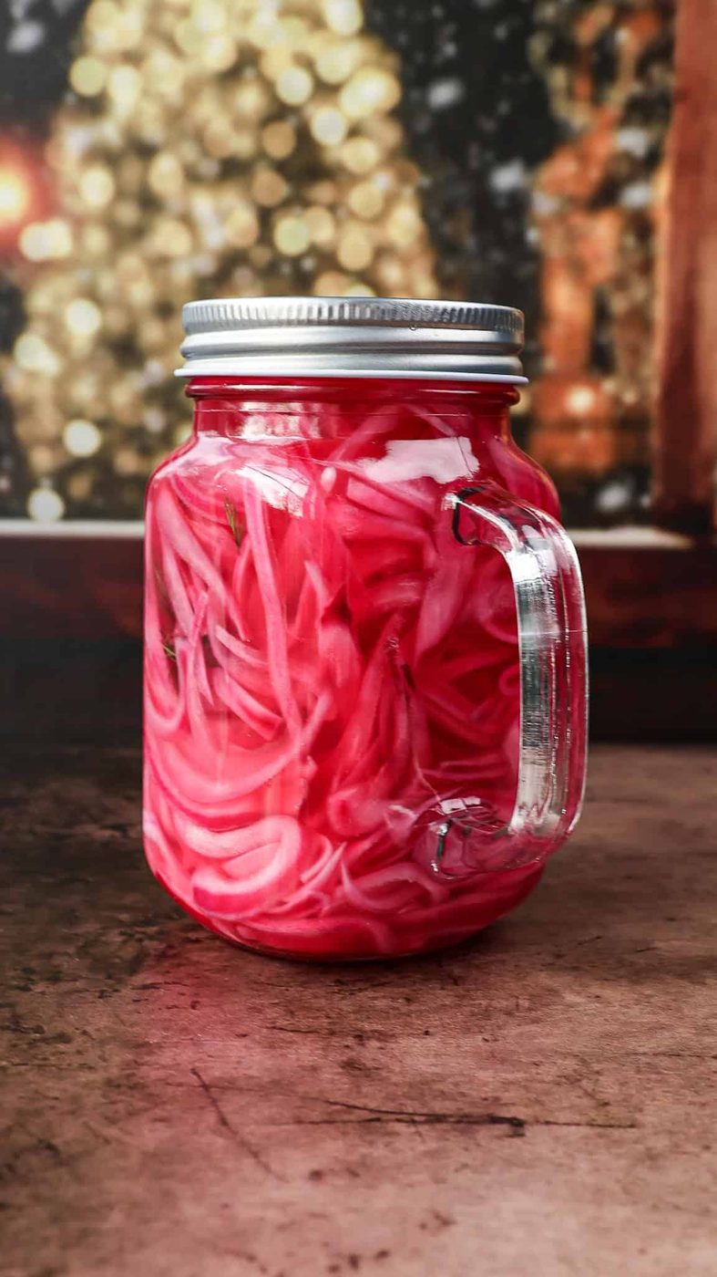 A glass jar with a handle, filled with bright pink pickled red onions, sits on a rustic surface with a blurred, festive background of warm lights.