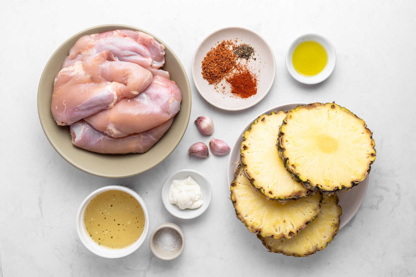 Flat lay of raw chicken thighs, sliced fresh pineapple, garlic cloves, assorted spices, olive oil, a bowl of marinade, a small dish of mayonnaise, and a dish of salt on a white surface.