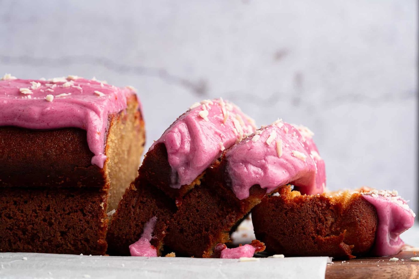 A loaf cake with vibrant pink frosting is partly sliced, revealing its moist interior. Crumbs and bits of frosting are scattered around on a light-colored surface, contrasting with the rustic background.