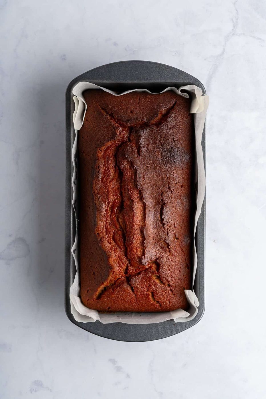 A freshly baked loaf of banana bread in a rectangular metal pan, lined with parchment paper, sits on a light marble surface. The bread's top is golden brown with a few cracks.