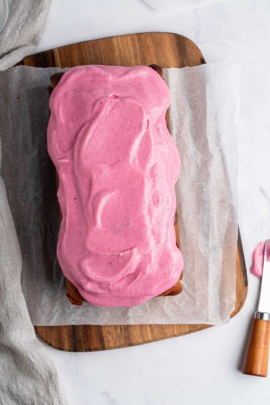 A rectangular loaf cake with bright pink frosting on a wooden board, partially covered with parchment paper. A knife with pink frosting is placed nearby on a white surface. A gray cloth is draped beside the cake.