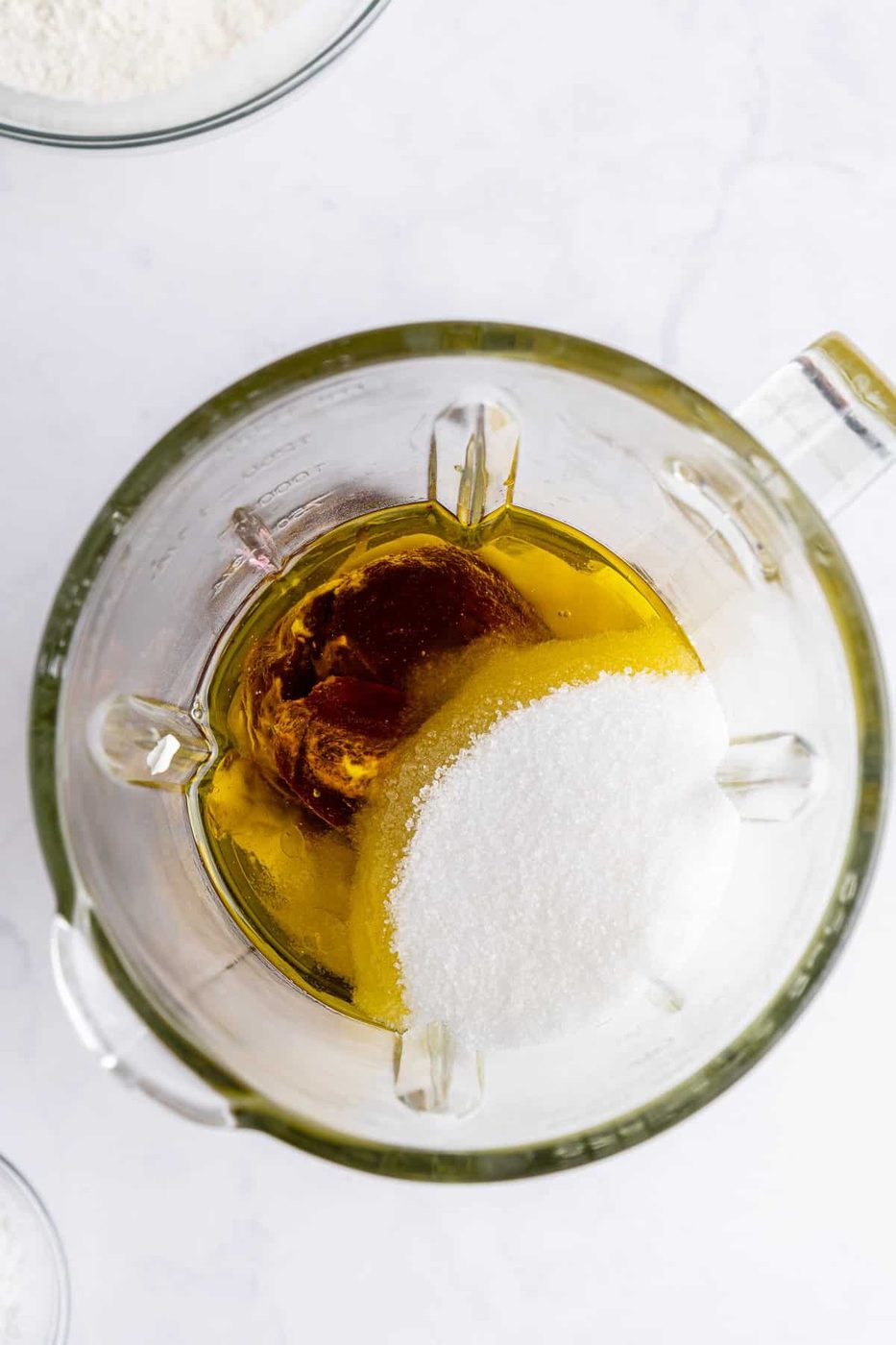 Top view of a blender pitcher containing oil, sugar, and vanilla extract on a white surface. The clear glass shows the ingredients before mixing.