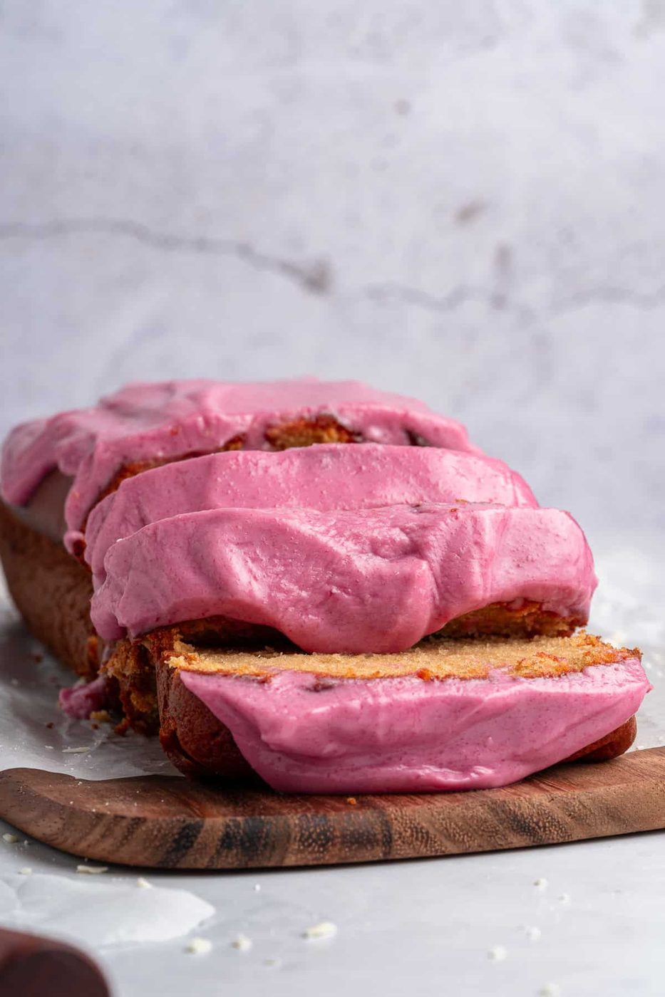 A sliced loaf cake topped with vibrant pink frosting sits on a wooden board against a light gray background. The cake appears moist and the frosting is generously spread, showcasing a rich and creamy texture.