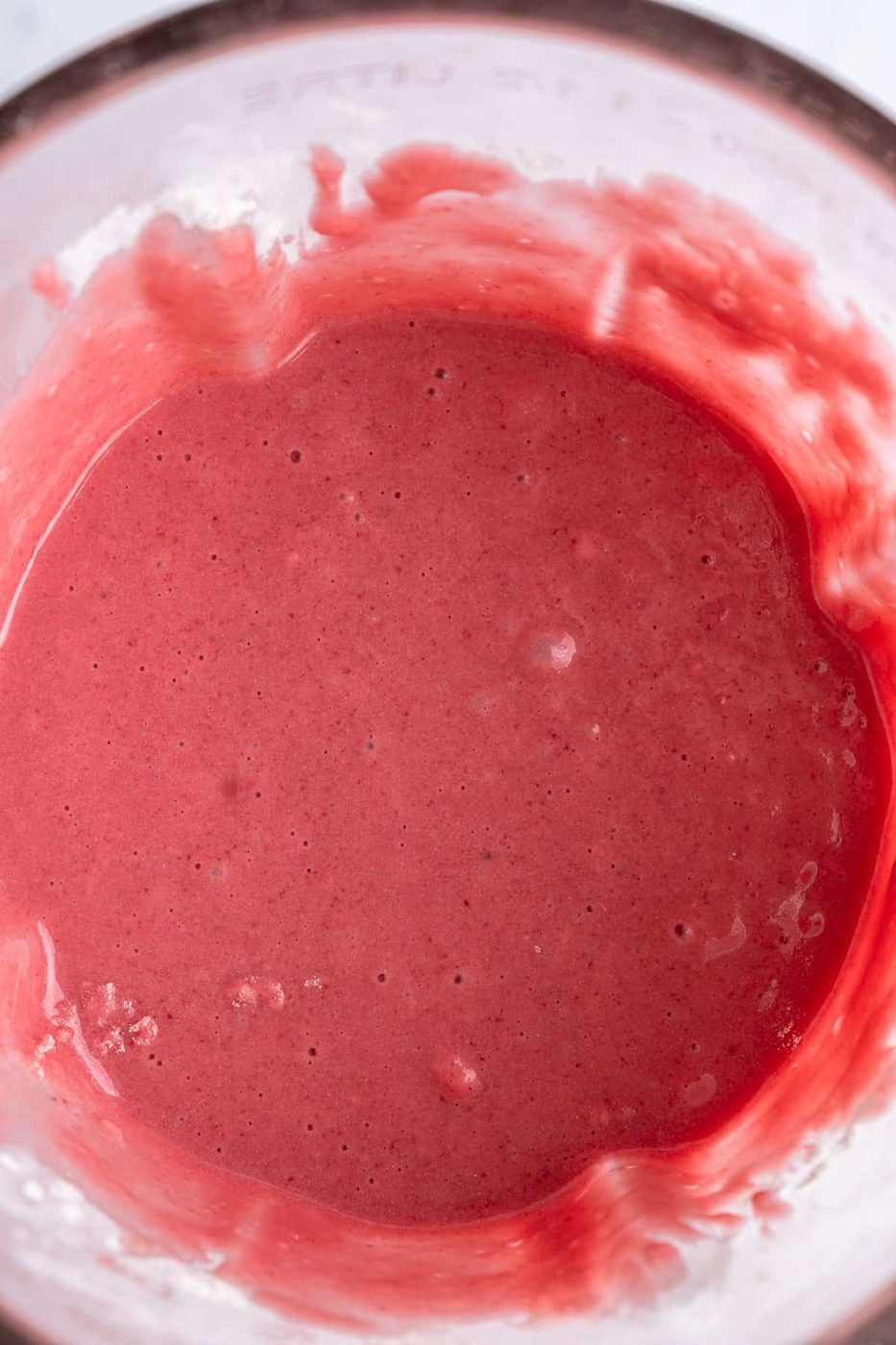 A glass filled with thick, smooth pink smoothie, featuring a creamy texture with small bubbles and an uneven surface. The smoothie is vibrant, suggesting a blend of fruits like berries, creating a refreshing and inviting appearance.