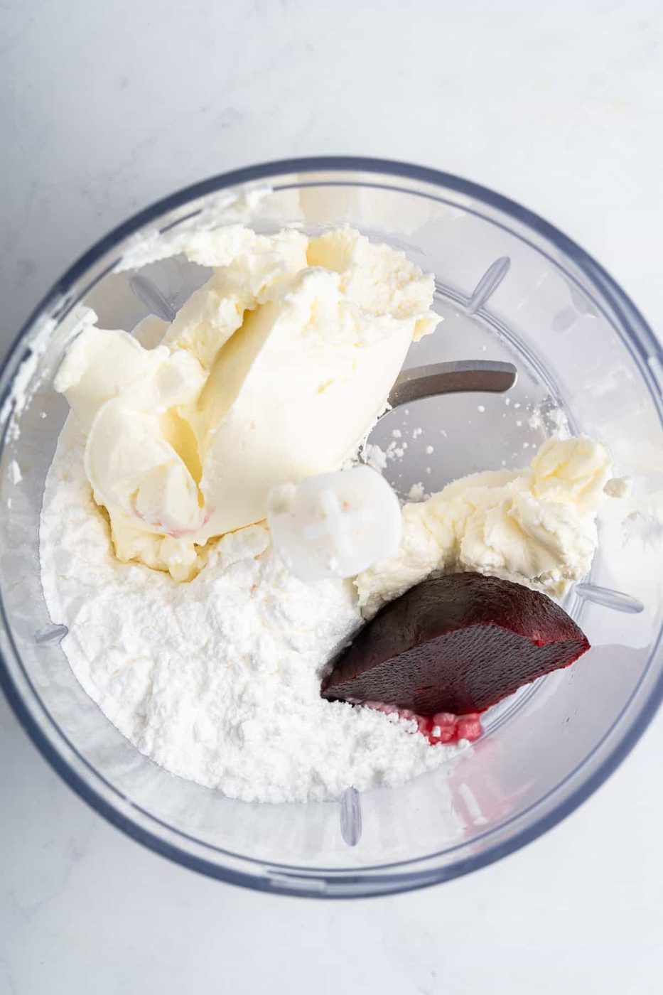 A food processor bowl containing cream cheese, powdered sugar, and a dollop of red food coloring or beet puree, ready to be mixed. The ingredients are arrayed on a light countertop.