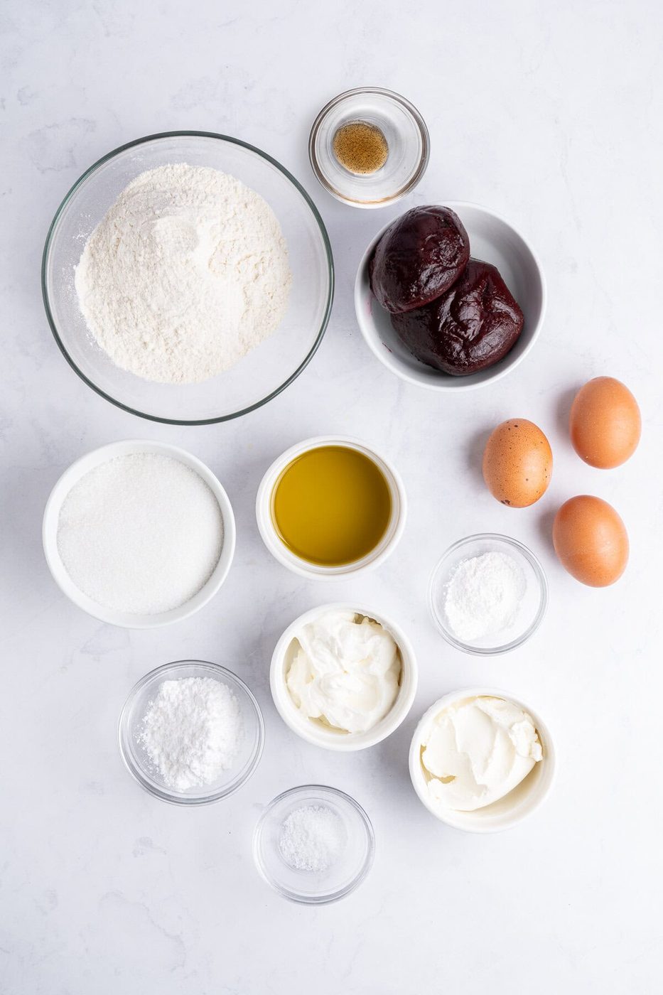 Top-down view of baking ingredients including flour, sugar, two beets, eggs, olive oil, baking powder, baking soda, yogurt, salt, and vanilla extract, all arranged in separate bowls on a white surface.