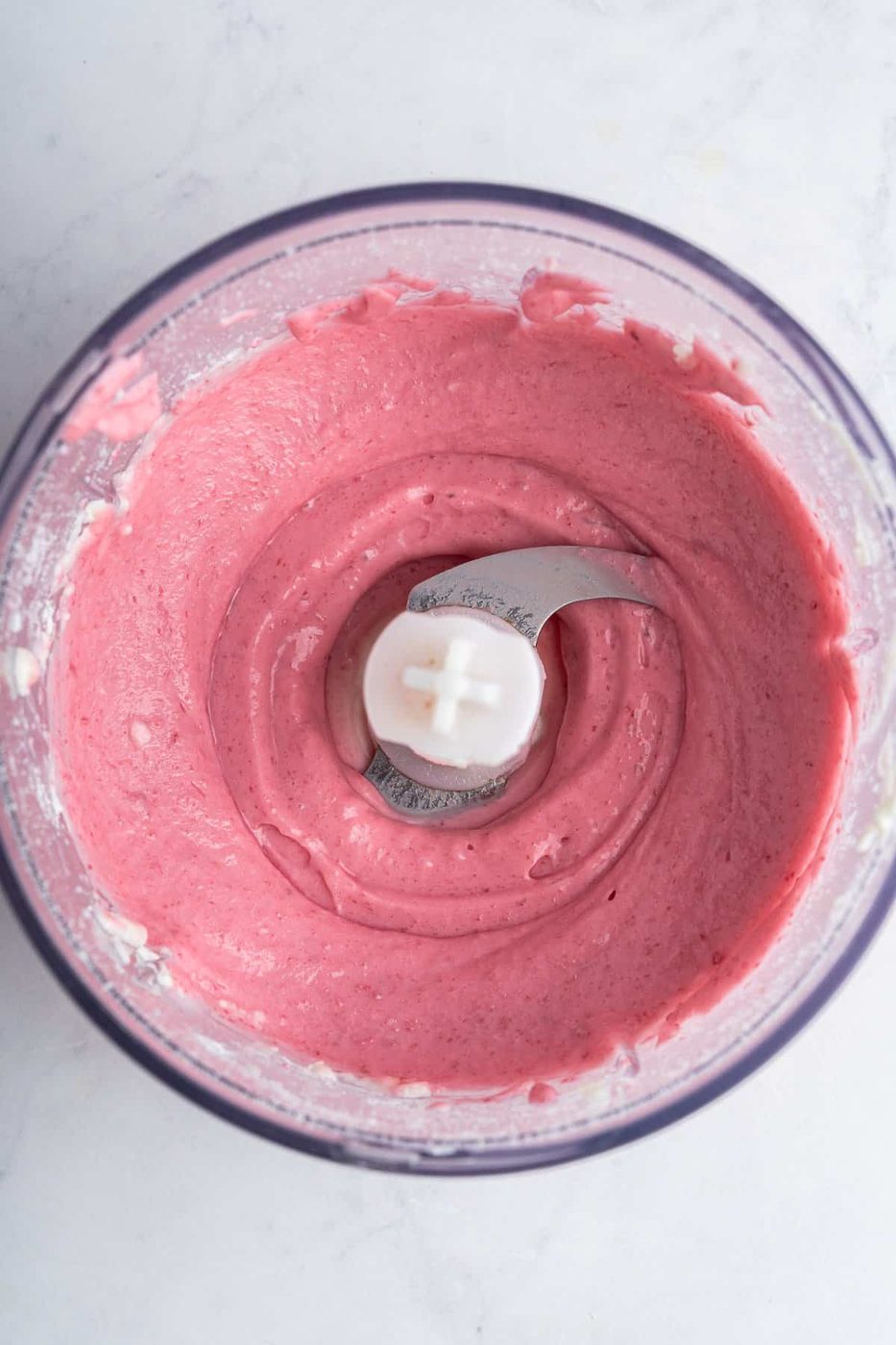 A top-down view of a food processor containing a smooth, blended pink mixture. The metal blade is visible in the center, resting within the creamy consistency. The background is a lightly textured white surface.